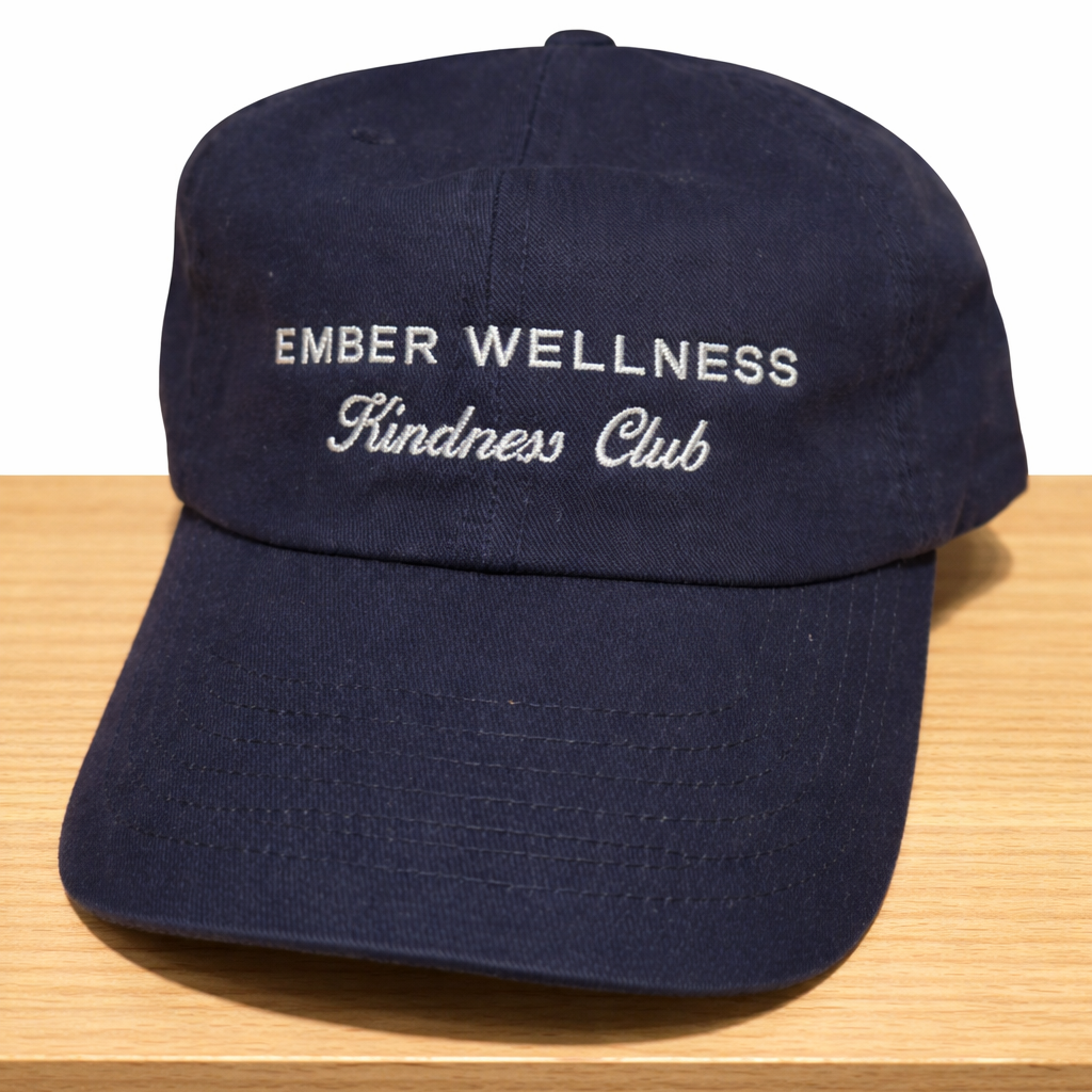Navy blue cap with 'Ember Wellness Kindness Club' text on a wooden surface.